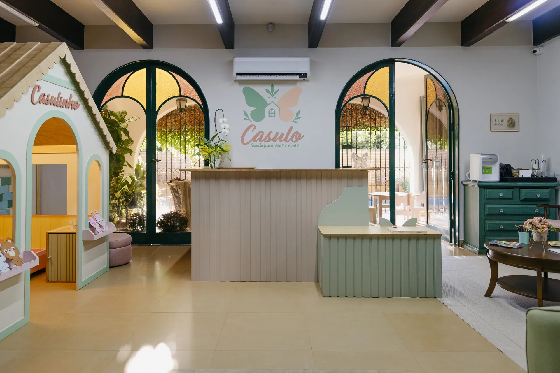 Recepção do Espaço Casulo, com portas em arco, balcão verde sálvia e a área infantil ao fundo